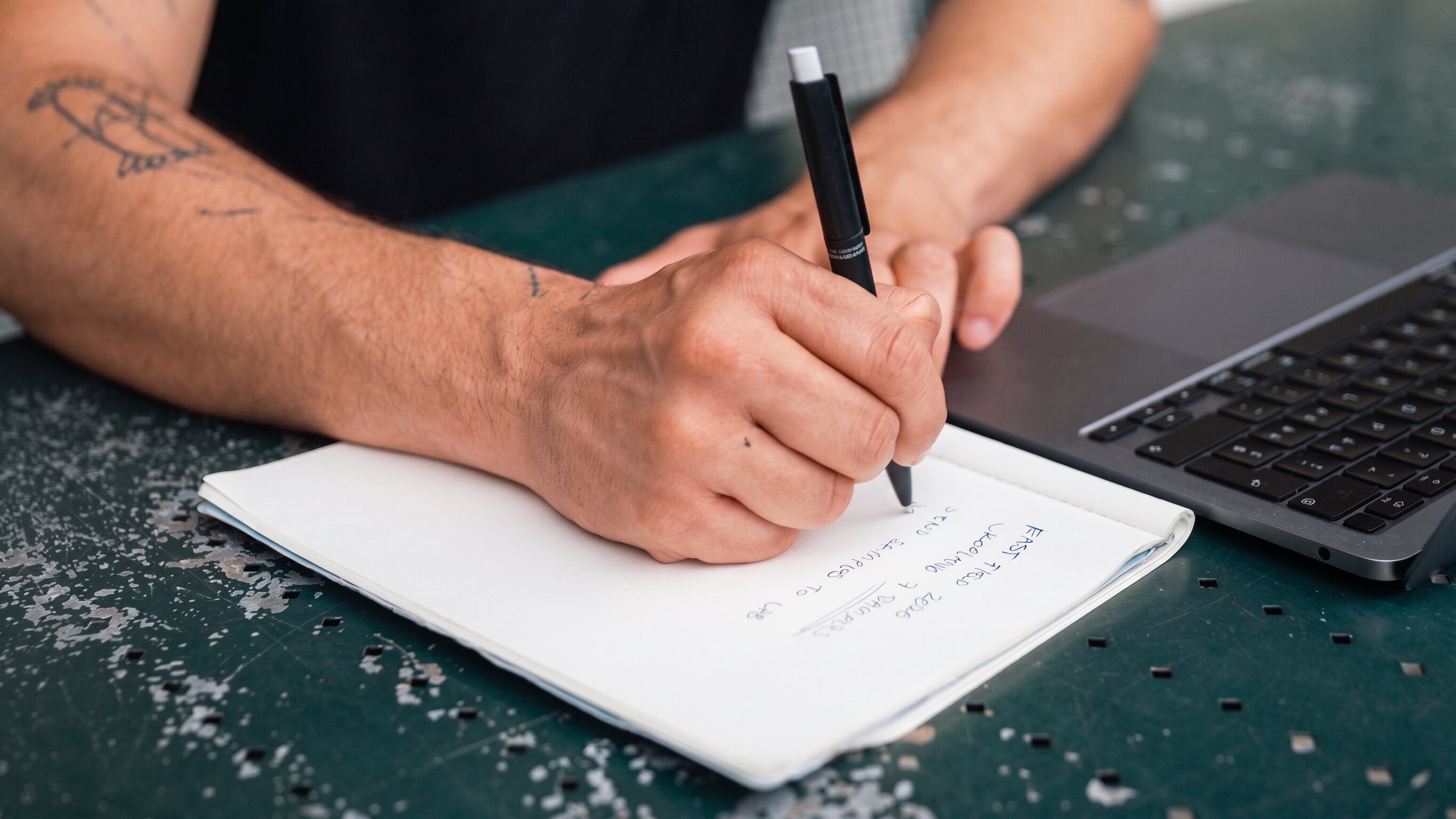 Hands writing field notes next to a laptop